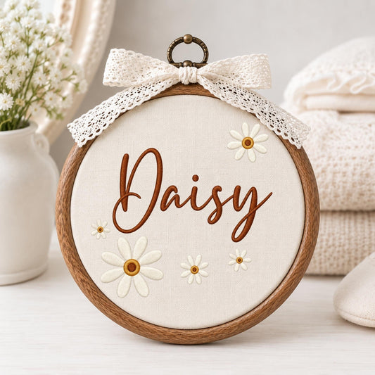 May include: A circular wooden embroidery hoop containing cream-colored fabric with the name Daisy embroidered in brown script. The design features several white daisy flowers with yellow centers and a decorative white lace bow at the top.