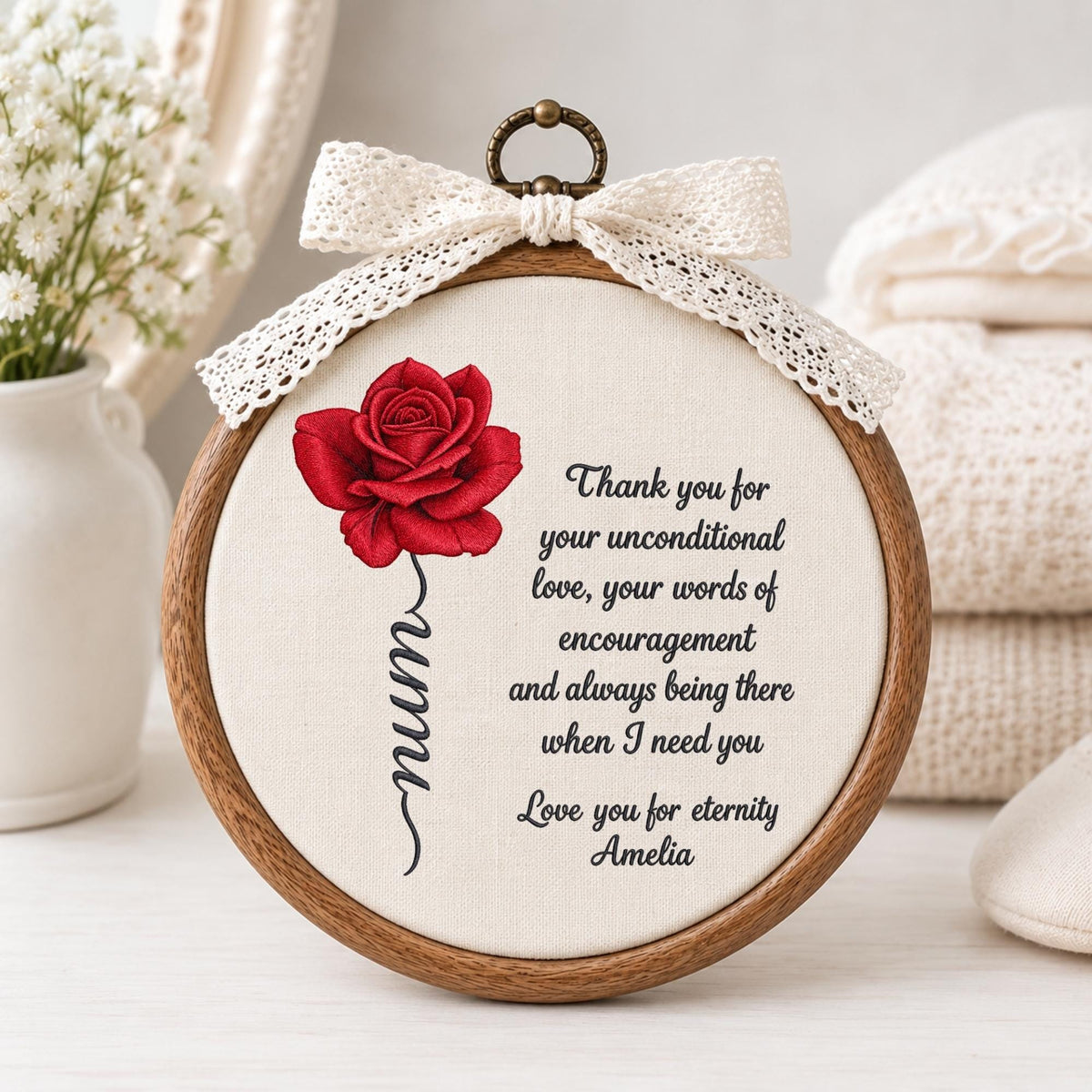 May include: A circular wooden embroidery hoop containing cream fabric with a red rose illustration, the word mum written in cursive, and black text reading Thank you for your unconditional love, your words of encouragement and always being there when I need you. Love you for eternity Amelia. A white lace bow is attached to the top.