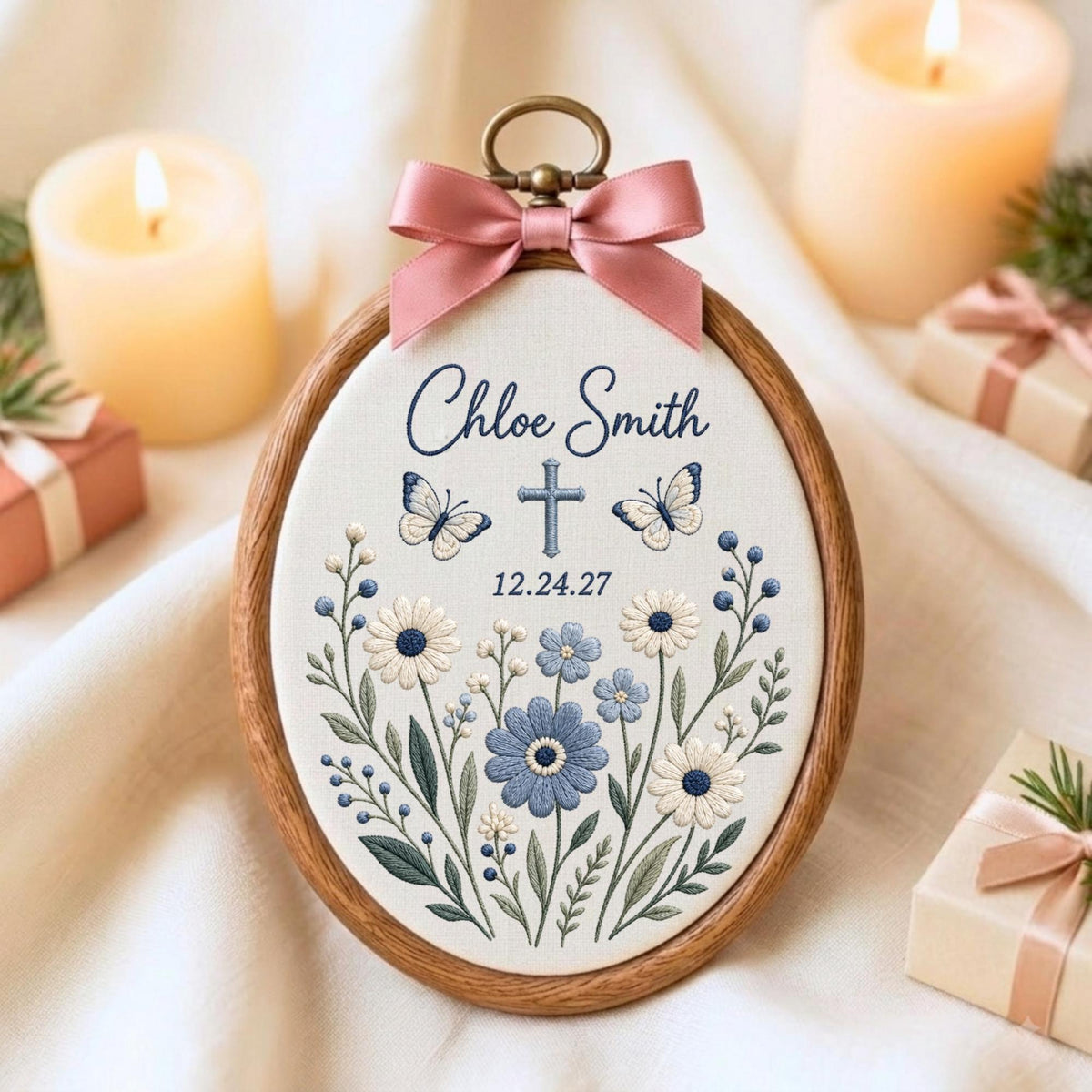May include: An oval wooden embroidery hoop containing white fabric with floral embroidery in shades of blue and white. The text reads Chloe Smith and 12.24.27, featuring a central cross and two butterflies. A pink satin ribbon bow is attached at the top.