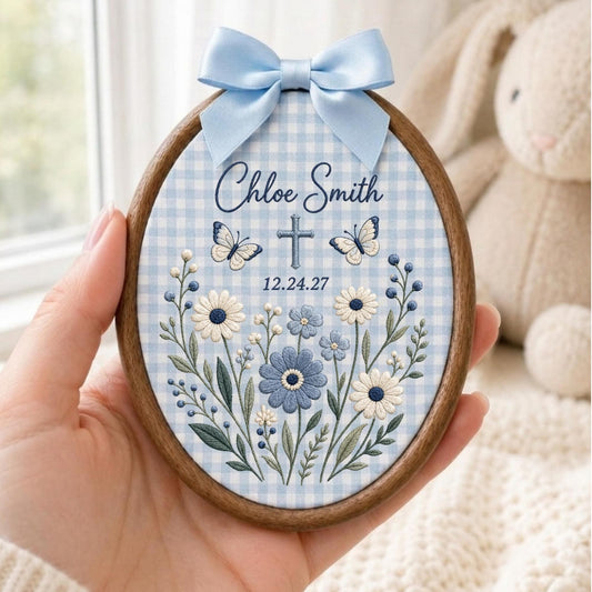 May include: An oval wooden embroidery hoop featuring light blue and white gingham fabric. The design includes embroidered white and blue flowers, two butterflies, a cross, and the text Chloe Smith 12.24.27. A light blue satin bow is attached at the top.