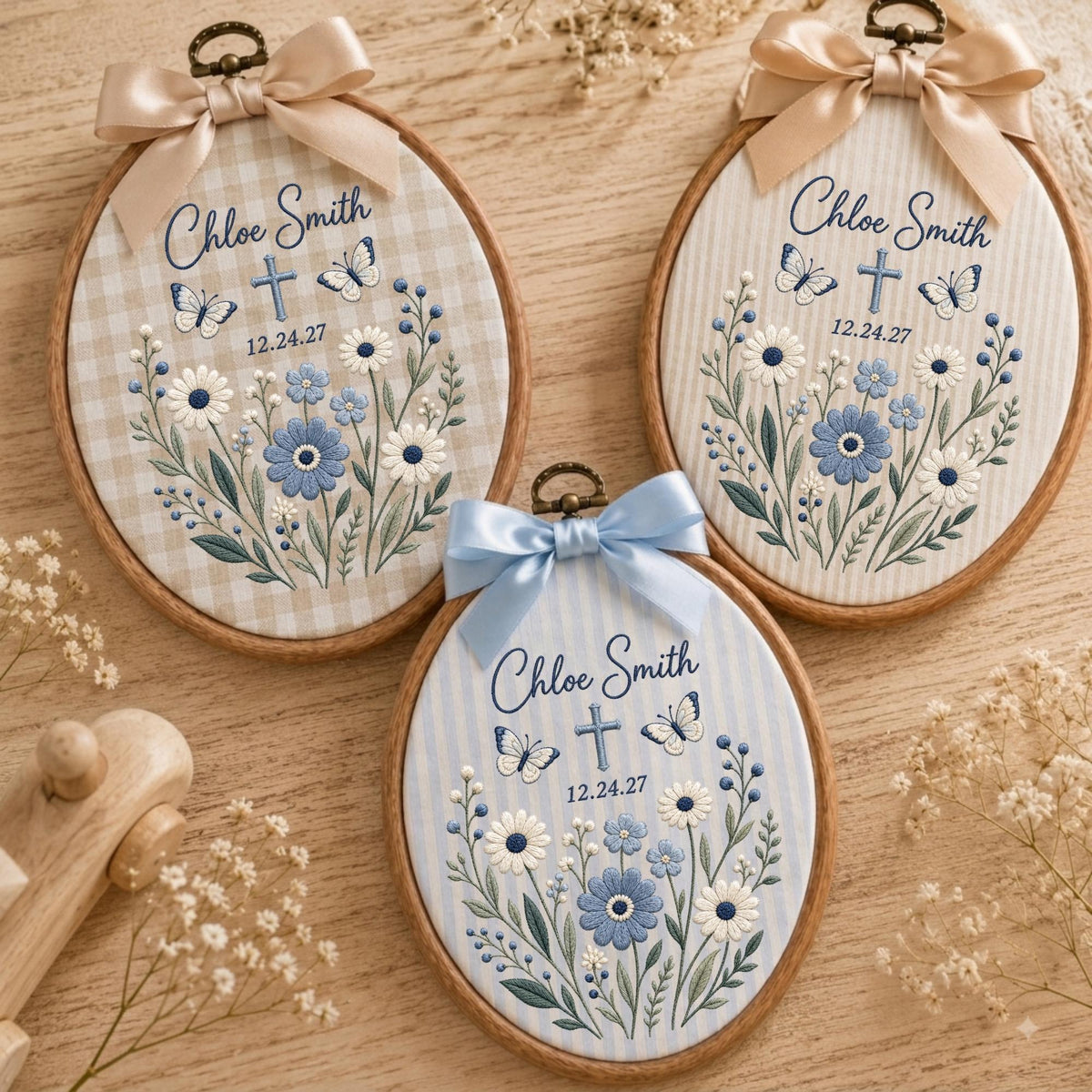 May include: Three oval wooden embroidery hoops featuring fabric backgrounds with embroidered blue and white flowers, butterflies, a cross, and the text Chloe Smith 12.24.27. Each hoop is topped with a decorative satin ribbon bow in beige or light blue.