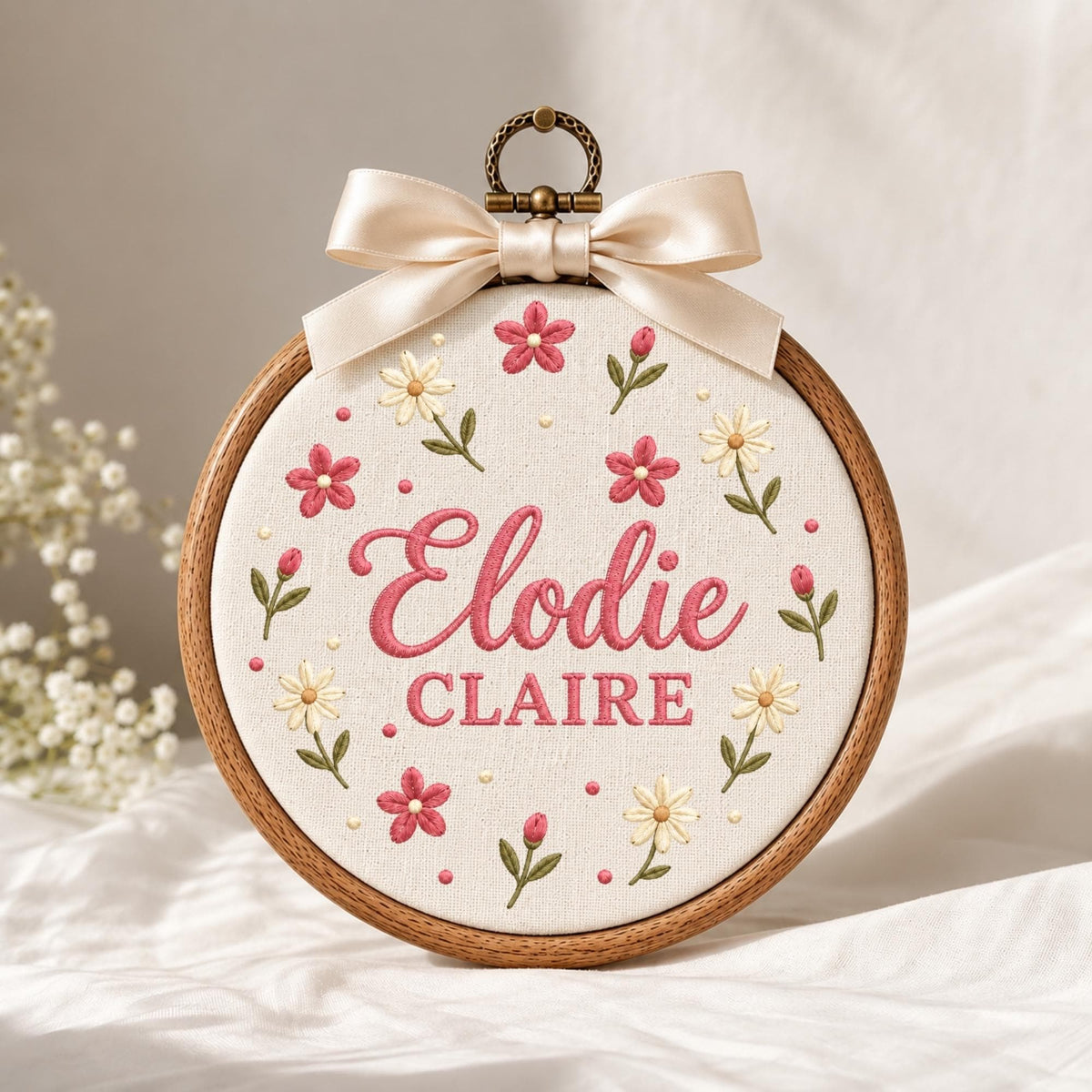 May include: A circular wooden embroidery hoop containing off-white fabric with the embroidered names Elodie CLAIRE in pink. The design features small pink and white flowers with green stems and a cream-colored satin ribbon bow at the top.