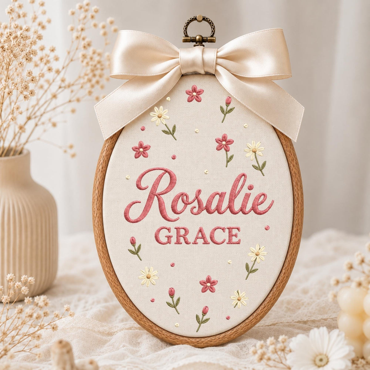 May include: An oval wooden embroidery hoop containing cream-colored fabric with the embroidered name Rosalie GRACE in pink and red lettering. The design features small embroidered pink and white flowers and a large cream satin bow at the top.