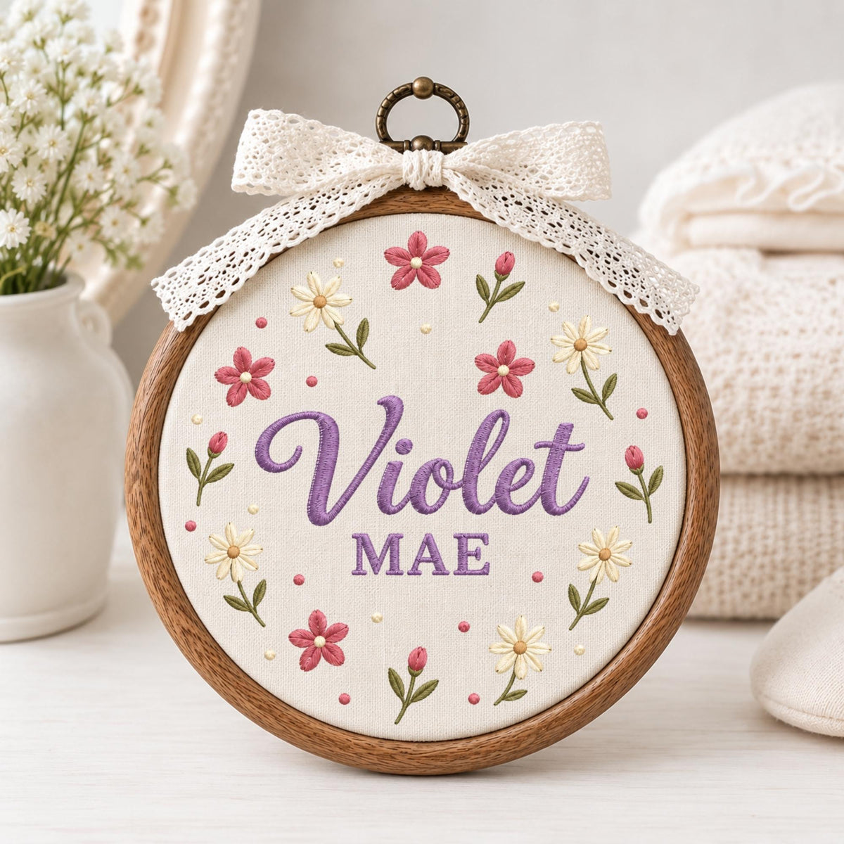 May include: A circular wooden embroidery hoop containing off-white fabric with the embroidered purple name Violet MAE in the center. The design features a floral wreath of pink and cream flowers with green stems and a cream lace bow on top.