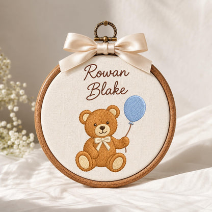 May include: A circular wooden embroidery hoop containing cream-colored fabric with the embroidered text Rowan Blake above a brown teddy bear holding a light blue balloon. A cream-colored satin ribbon bow is attached to the top metal hanger.