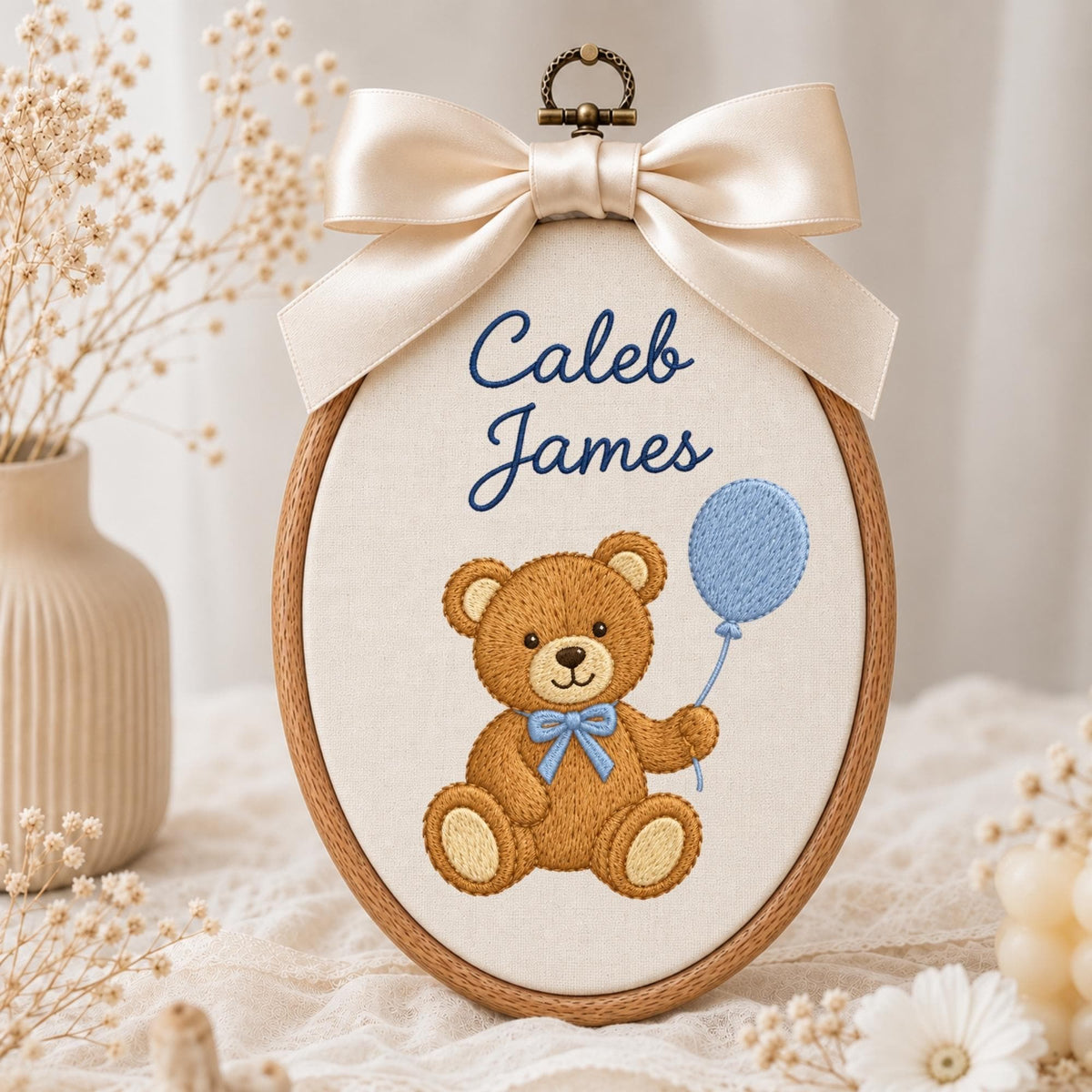 May include: An oval embroidery hoop featuring a cream-colored fabric background with the embroidered name Caleb James in dark blue script. Below the text is a brown teddy bear with a light blue bow tie holding a light blue balloon.
