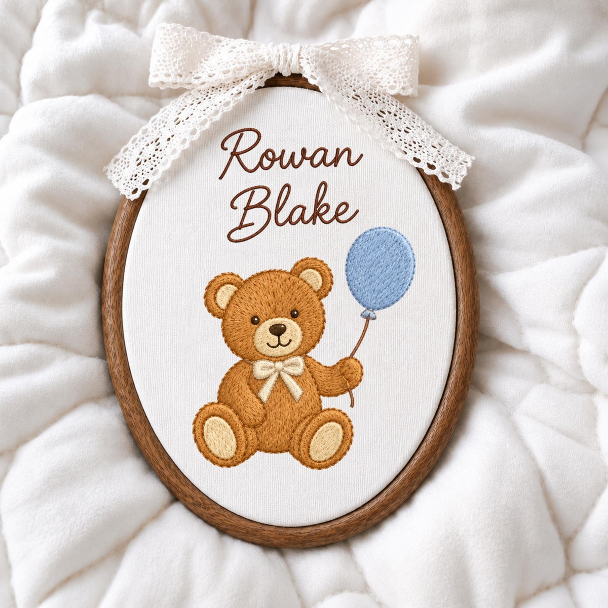 May include: An oval wooden embroidery hoop containing white fabric with the embroidered text Rowan Blake in brown. Below the text is a brown teddy bear with a cream bow tie holding a light blue balloon. A white lace bow is attached to the top.