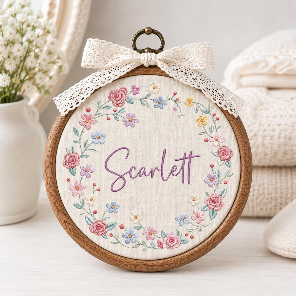 May include: A circular wooden embroidery hoop containing off-white fabric with a floral wreath design embroidered in pink, purple, blue, and yellow threads. The name Scarlett is stitched in purple script in the center, topped with a cream lace bow.