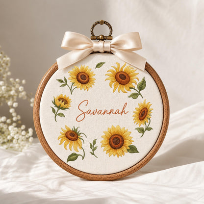May include: A circular wooden embroidery hoop containing off-white fabric with a sunflower wreath design and the name Savannah embroidered in brown script. A cream-colored satin ribbon bow is attached to the top metal hanging loop.