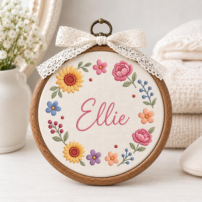 May include: A circular wooden embroidery hoop containing white fabric with the name Ellie stitched in pink cursive script. The name is surrounded by a colorful floral wreath featuring yellow sunflowers, pink roses, and small blue and orange flowers.