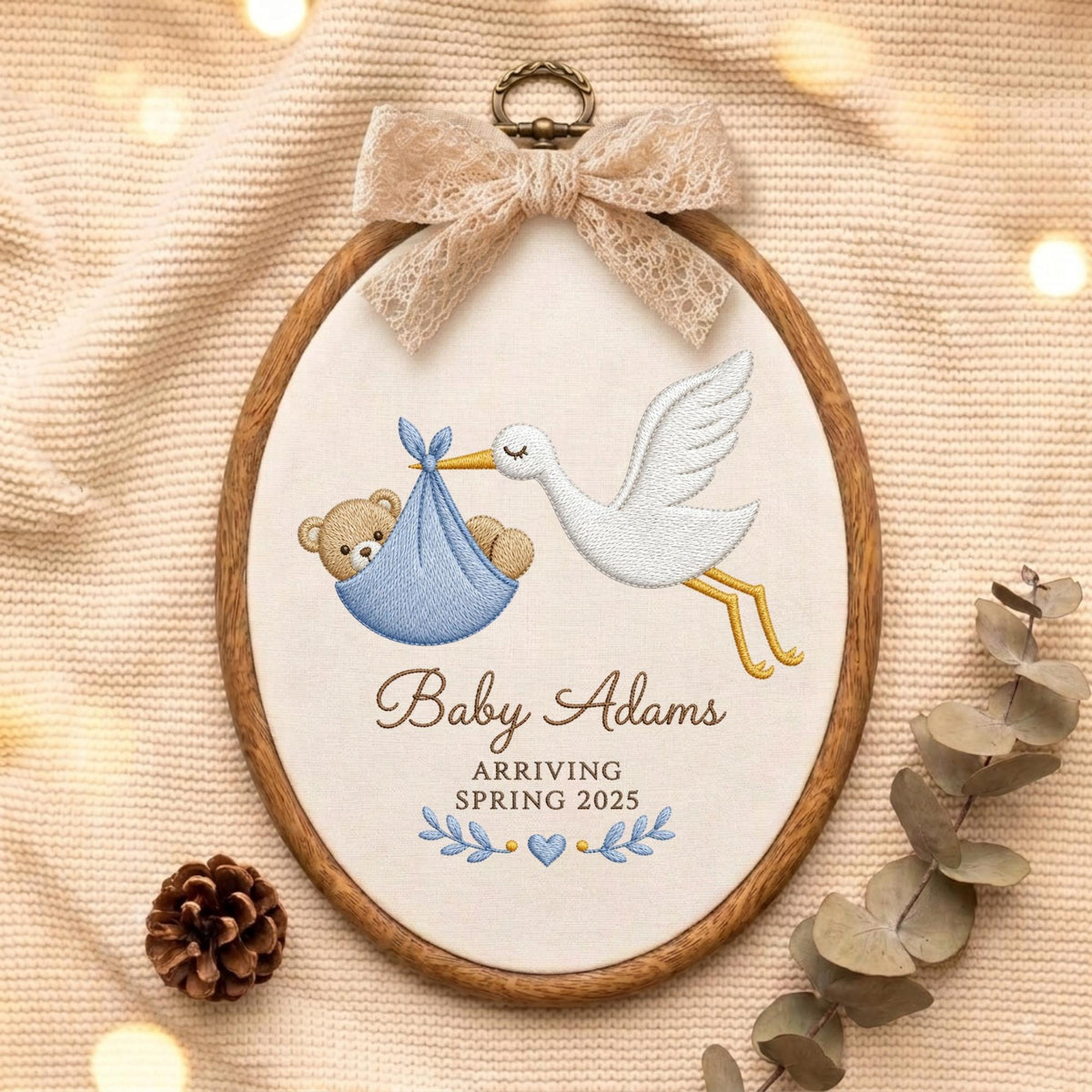 May include: An oval wooden embroidery hoop containing white fabric with a stitched design of a white stork carrying a blue bundle with a teddy bear inside. The text reads Baby Adams ARRIVING SPRING 2025. A beige lace bow is attached to the top.