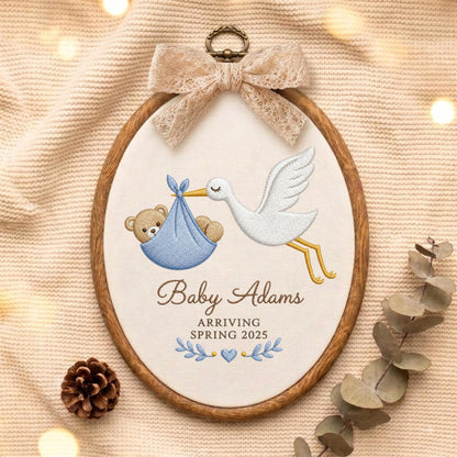 May include: An oval wooden embroidery hoop containing white fabric with a stitched design of a white stork carrying a blue bundle with a teddy bear inside. The text reads Baby Adams ARRIVING SPRING 2025. A beige lace bow is attached to the top.