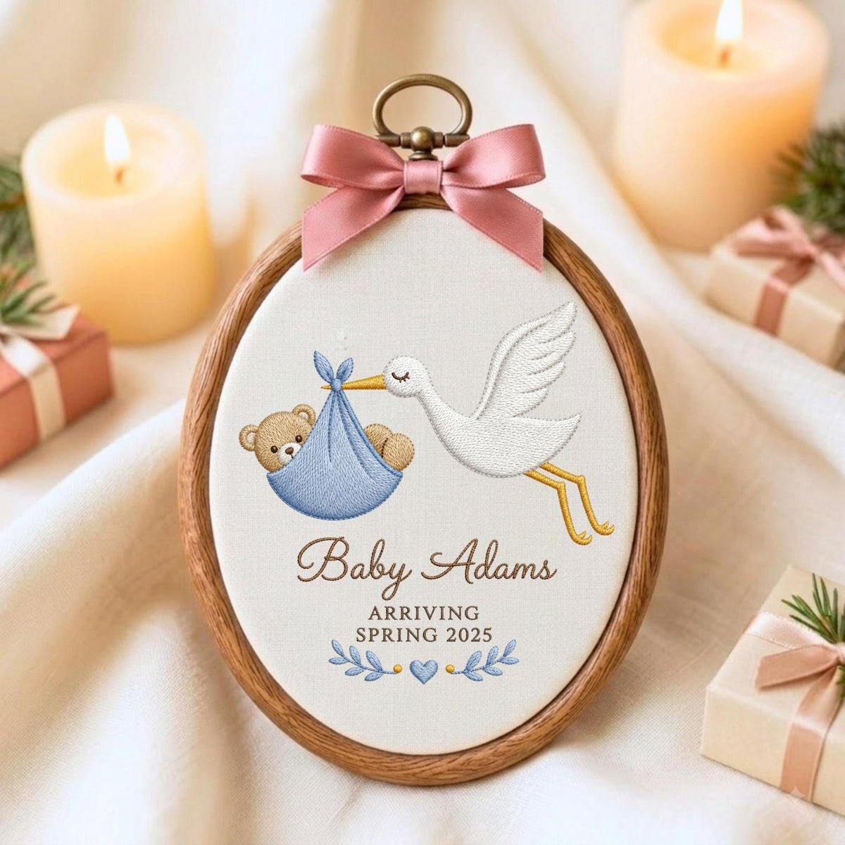 May include: An oval wooden embroidery hoop containing white fabric with an embroidered white stork carrying a blue bundle with a brown teddy bear inside. The text reads Baby Adams ARRIVING SPRING 2025 with blue leaves and a heart. A pink satin bow is attached.