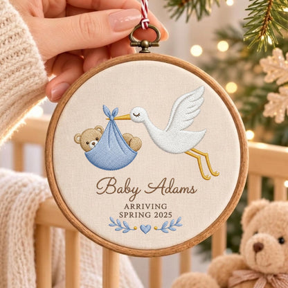 May include: A round wooden embroidery hoop featuring a white fabric center with an embroidered white stork carrying a blue bundle containing a brown teddy bear. The text reads Baby Adams ARRIVING SPRING 2025 above blue leaf and heart accents.