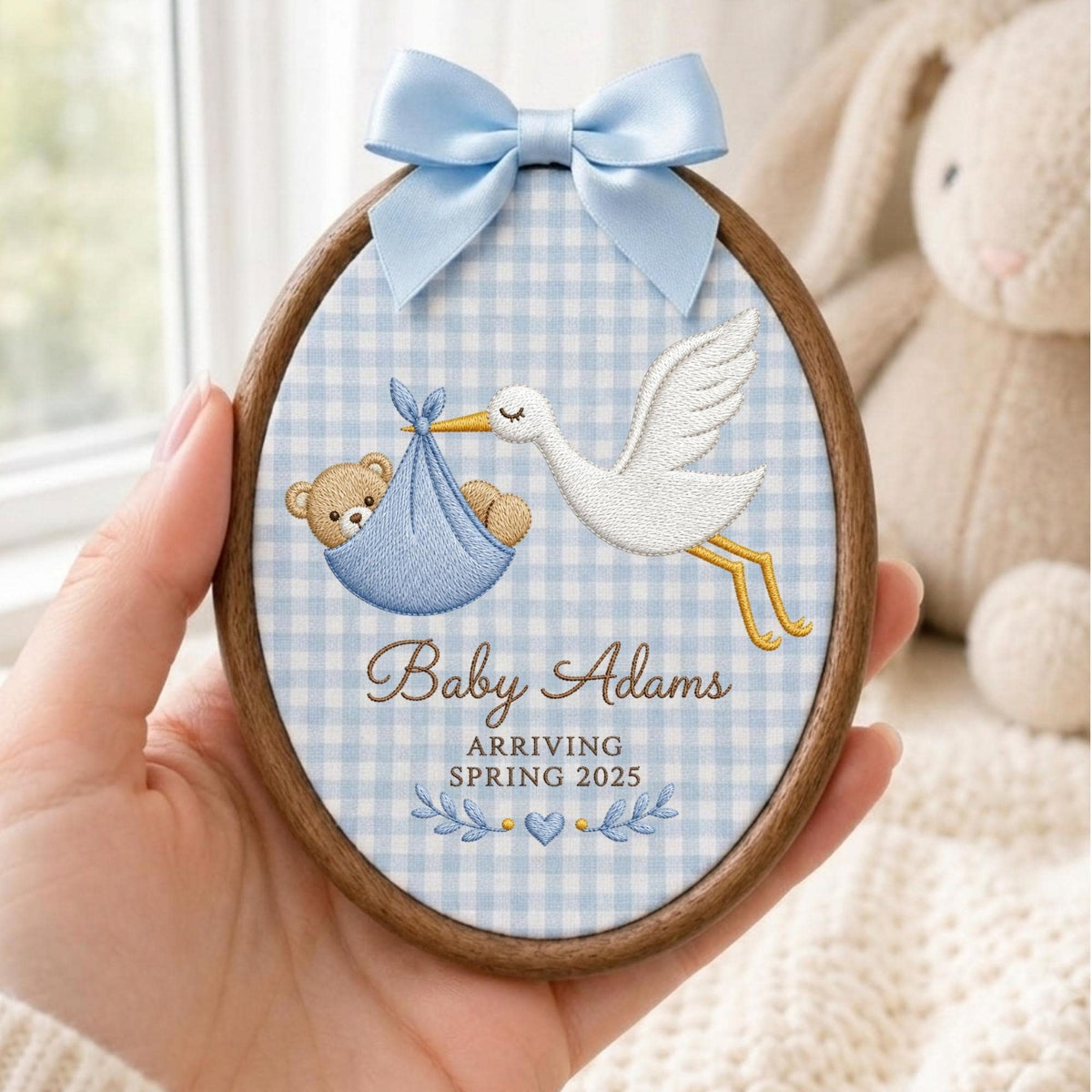 May include: An oval wooden embroidery hoop featuring a light blue and white gingham fabric background. The embroidery depicts a white stork carrying a brown teddy bear in a blue bundle. Text reads Baby Adams ARRIVING SPRING 2025 with a blue bow.