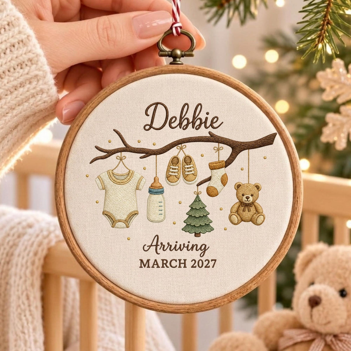 May include: A round wooden embroidery hoop featuring a cream-colored fabric with an embroidered brown tree branch. Hanging from the branch are a baby onesie, a bottle, shoes, a sock, a small tree, and a teddy bear. Text reads Debbie Arriving MARCH 2027.