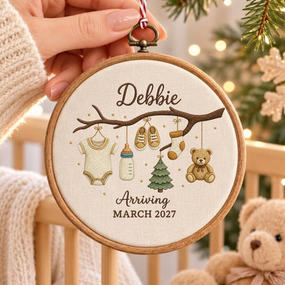 May include: A round wooden embroidery hoop featuring a cream-colored fabric with an embroidered brown tree branch. Hanging from the branch are a baby onesie, a bottle, shoes, a sock, a small tree, and a teddy bear. Text reads Debbie Arriving MARCH 2027.