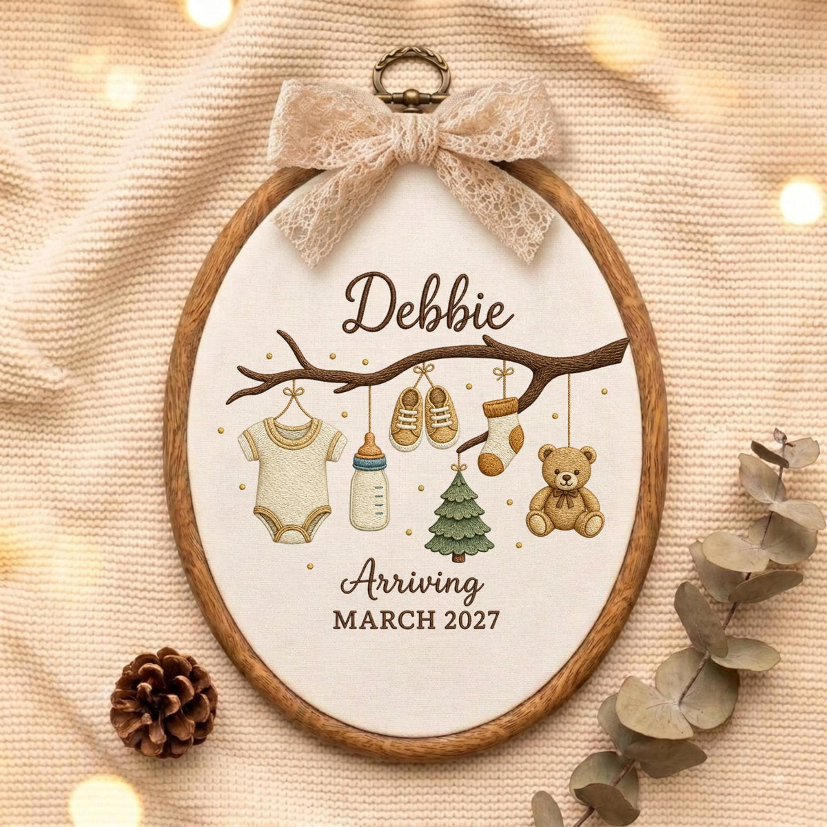 May include: An oval wooden embroidery hoop containing fabric with an embroidered branch. Hanging from the branch are a onesie, baby bottle, shoes, sock, Christmas tree, and teddy bear. Text reads Debbie Arriving MARCH 2027. A lace bow is at the top.