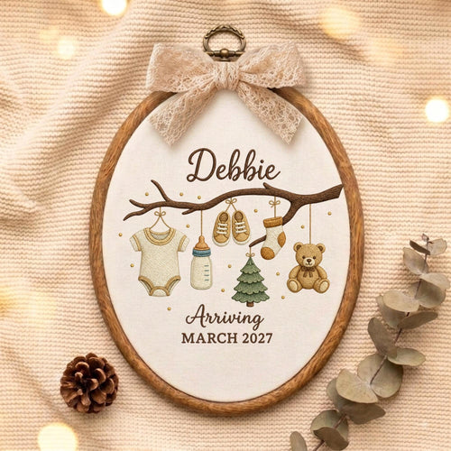May include: An oval wooden embroidery hoop containing fabric with an embroidered branch. Hanging from the branch are a onesie, baby bottle, shoes, sock, Christmas tree, and teddy bear. Text reads Debbie Arriving MARCH 2027. A lace bow is at the top.