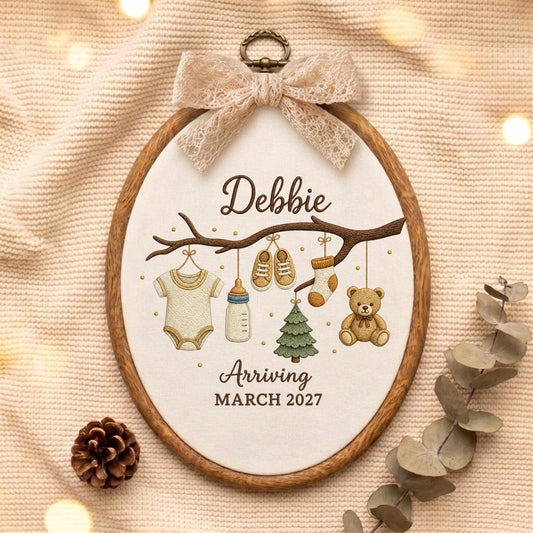 May include: An oval wooden embroidery hoop containing fabric with an embroidered branch. Hanging from the branch are a onesie, baby bottle, shoes, sock, Christmas tree, and teddy bear. Text reads Debbie Arriving MARCH 2027. A lace bow is at the top.