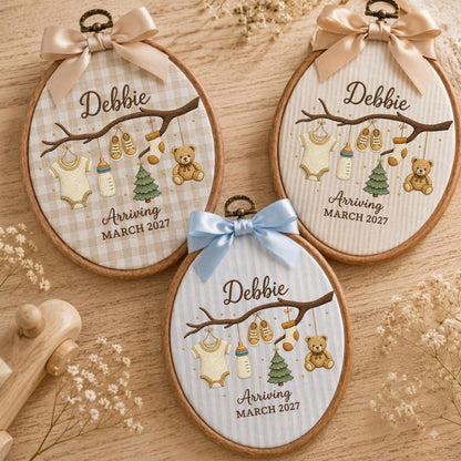 May include: Three oval wooden embroidery hoops featuring fabric with baby-themed illustrations of a onesie, shoes, socks, a bottle, a tree, and a teddy bear hanging from a branch. Each hoop displays the name Debbie and Arriving March 2027.