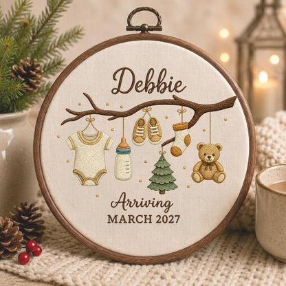May include: A circular wooden embroidery hoop containing fabric with a stitched design of a tree branch. Hanging from the branch are a baby onesie, a bottle, shoes, a sock, a tree, and a teddy bear. Text reads: Debbie Arriving MARCH 2027.