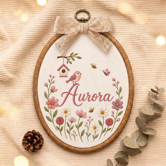 May include: An oval wooden embroidery hoop containing white fabric with a stitched design of the name Aurora in pink script, a pink bird, a birdhouse, bees, a butterfly, and various colorful flowers, topped with a beige lace bow.