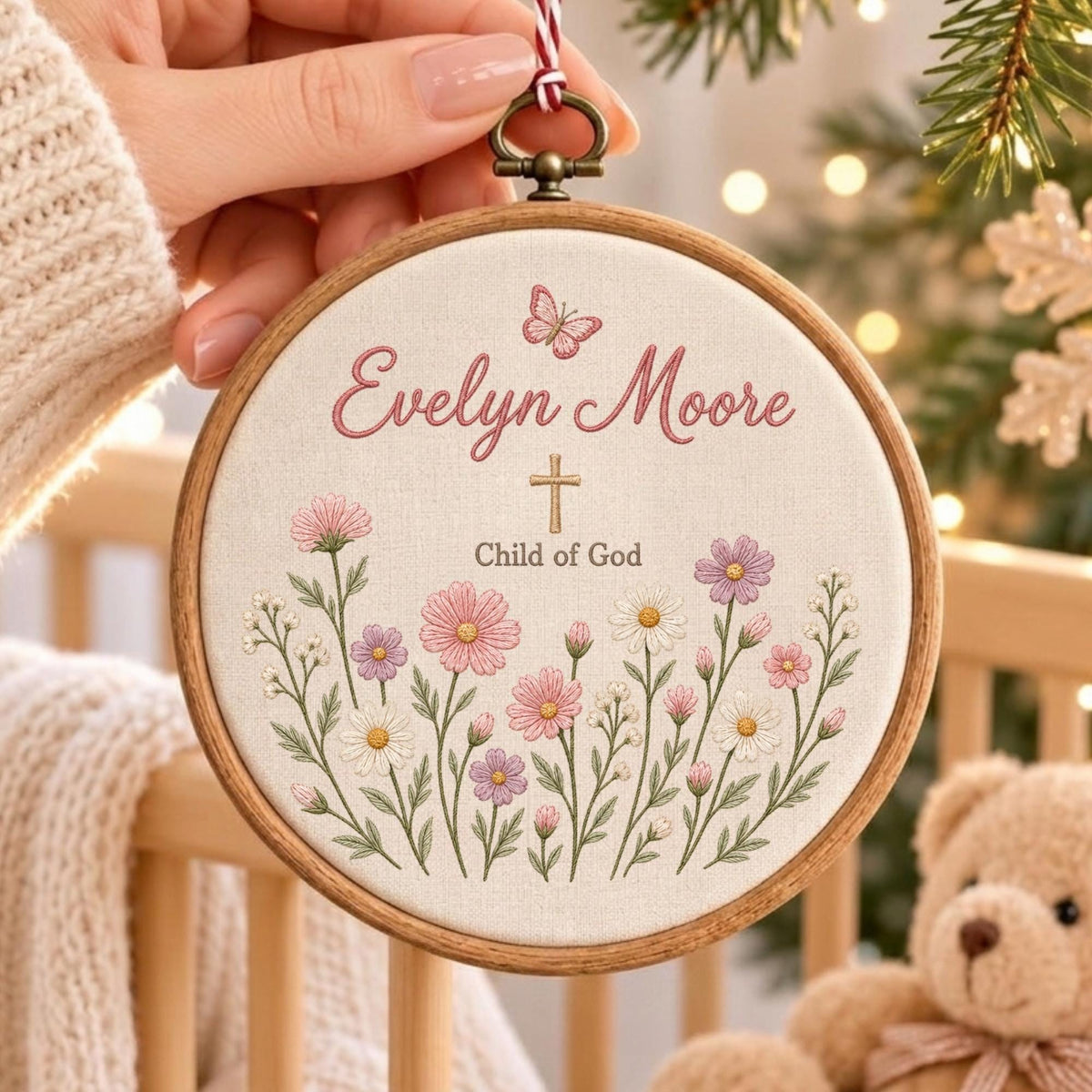 May include: A circular wooden embroidery hoop containing off-white fabric with floral embroidery in pink, white, and purple. The text reads Evelyn Moore and Child of God, centered with a small cross and a butterfly above the name in pink script.