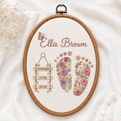 May include: An oval embroidery hoop containing fabric with stitched floral footprints, a butterfly, and the text Ella Brown, 12-18-2026, 2.9 KG, and 55 CM. The design features pink, purple, and yellow flowers arranged in the shape of two feet.