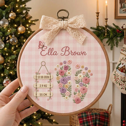 May include: A circular wooden embroidery hoop featuring a light pink gingham fabric background. The design includes the name Ella Brown, a butterfly, two floral-patterned baby footprints, and three hanging wooden signs with 12-18-2026, 2.9 KG, and 55 CM. A cream lace bow is attached to the top.