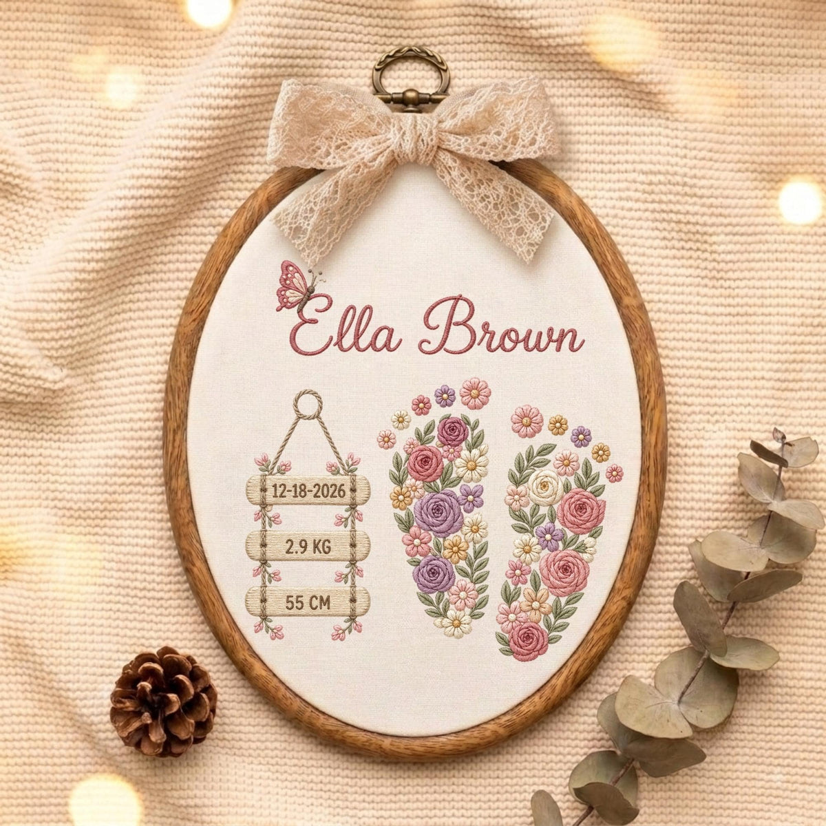 May include: An oval wooden embroidery hoop containing white fabric with a lace bow at the top. The embroidery features the name Ella Brown, a butterfly, two floral footprints, and three hanging signs displaying 12-18-2026, 2.9 KG, and 55 CM.