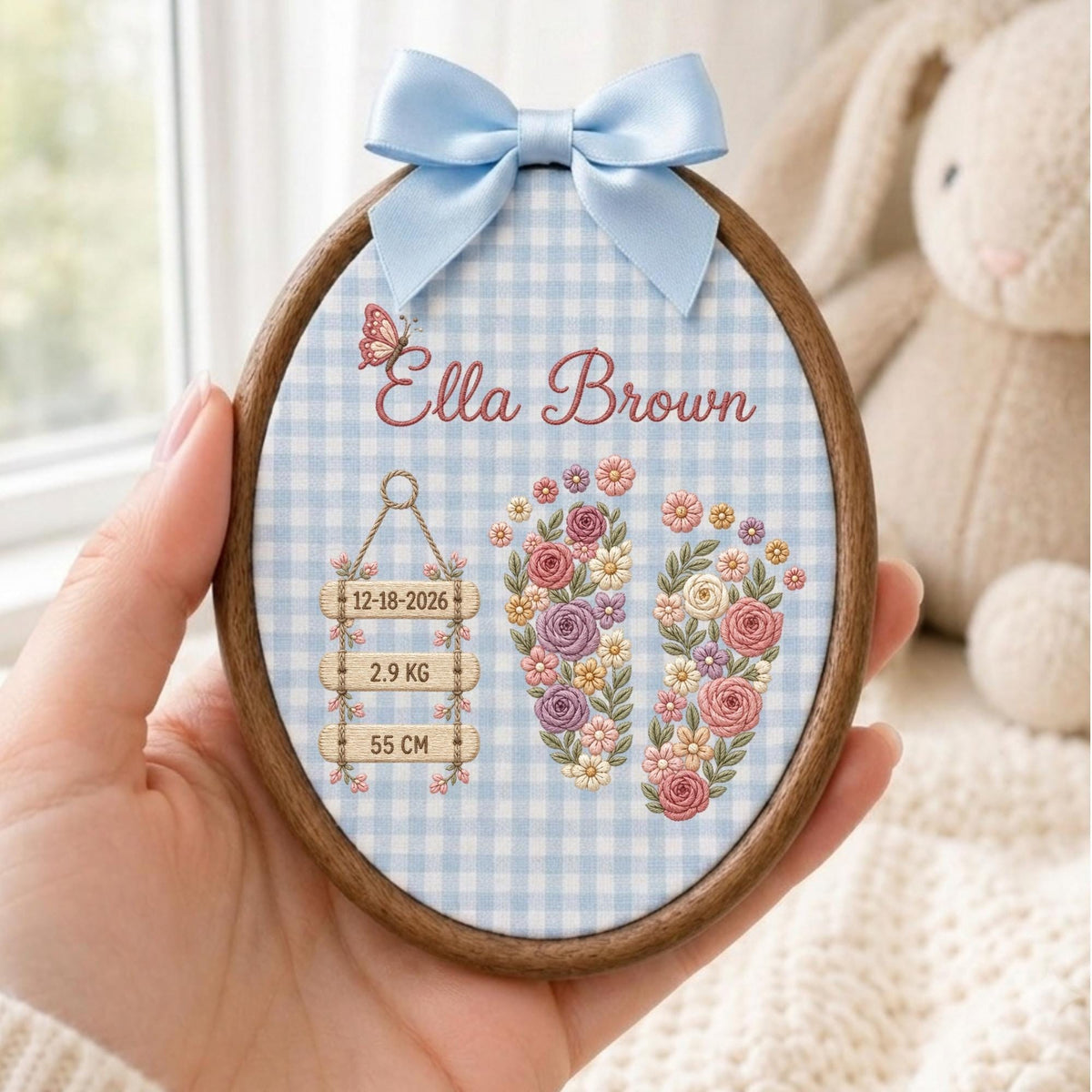 May include: An oval wooden embroidery hoop featuring a light blue and white gingham fabric background. The design includes the name Ella Brown, a butterfly, and two floral-filled baby footprints. Three wooden signs display 12-18-2026, 2.9 KG, and 55 CM. A light blue satin bow is attached to the top.