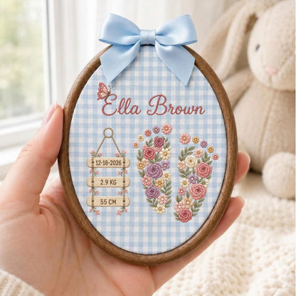 May include: An oval wooden embroidery hoop featuring a light blue and white gingham fabric background. The design includes the name Ella Brown, a butterfly, and two floral-filled baby footprints. Three wooden signs display 12-18-2026, 2.9 KG, and 55 CM. A light blue satin bow is attached to the top.