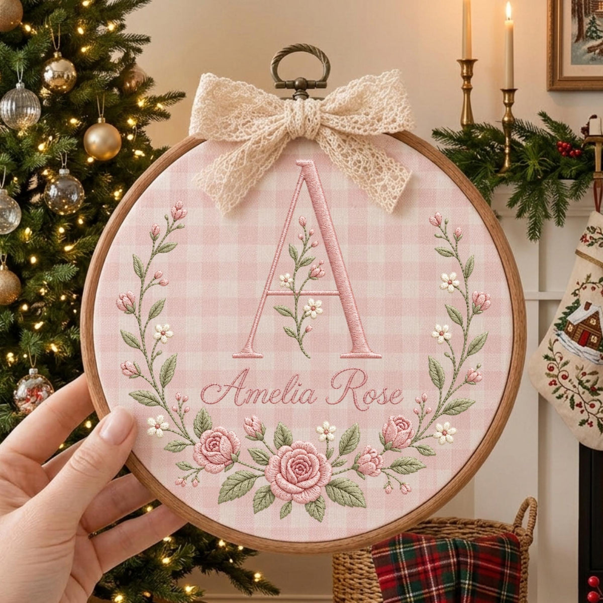 May include: A circular wooden embroidery hoop containing pink and white gingham fabric. The fabric features an embroidered large letter A, the name Amelia Rose in cursive script, and a floral wreath of pink roses and green leaves, topped with a lace bow.