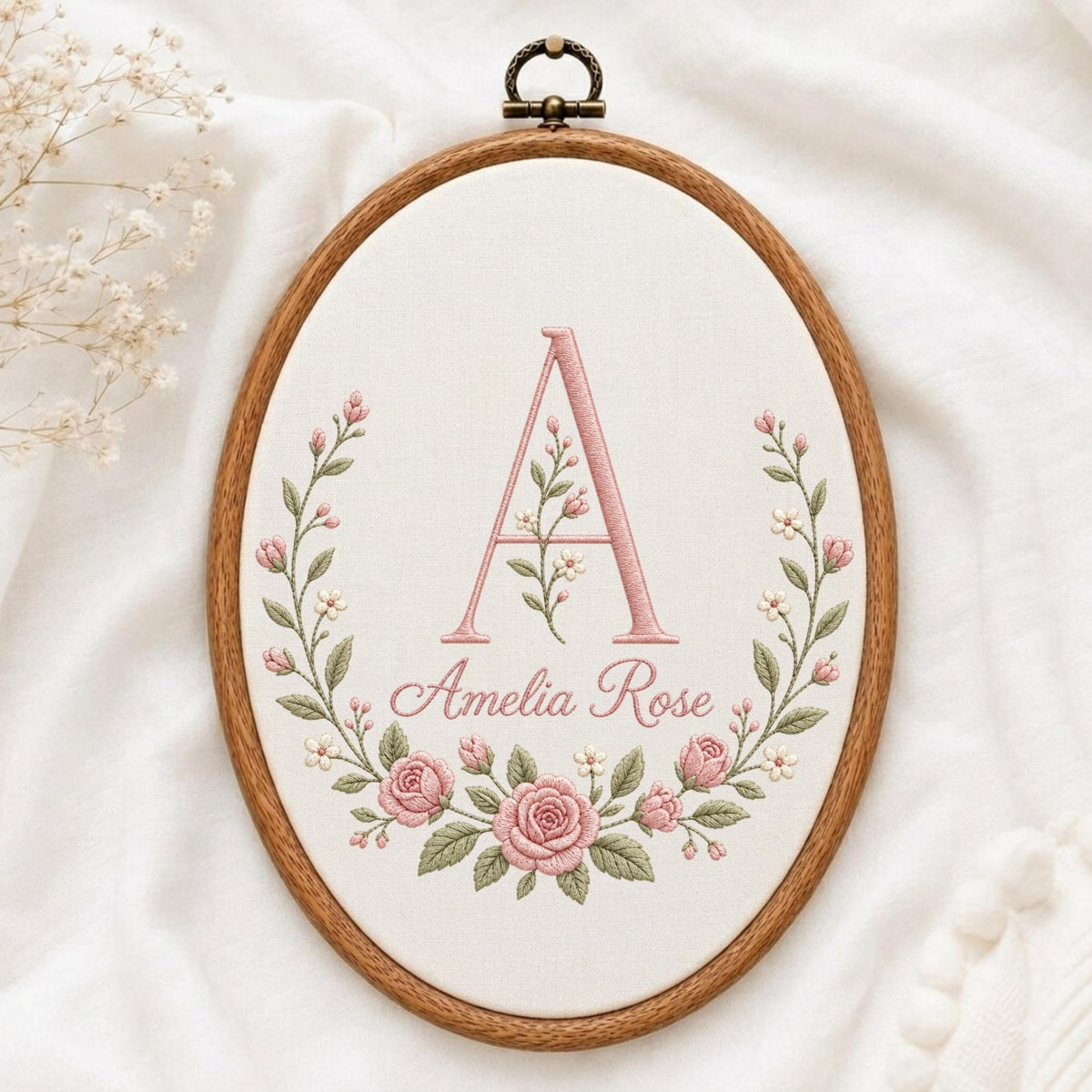 May include: An oval wooden embroidery hoop containing white fabric with a hand-stitched design. The embroidery features a large pink letter A, the name Amelia Rose in cursive script, and a floral wreath of pink roses and green leaves.