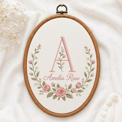 May include: An oval wooden embroidery hoop containing white fabric with a hand-stitched design. The embroidery features a large pink letter A, the name Amelia Rose in cursive script, and a floral wreath of pink roses and green leaves.