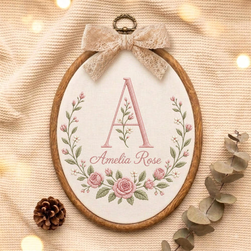 May include: An oval wooden embroidery hoop containing off-white fabric with a floral wreath design in pink and green. The center features a large letter A and the name Amelia Rose in pink script. A beige lace bow is attached to the top.