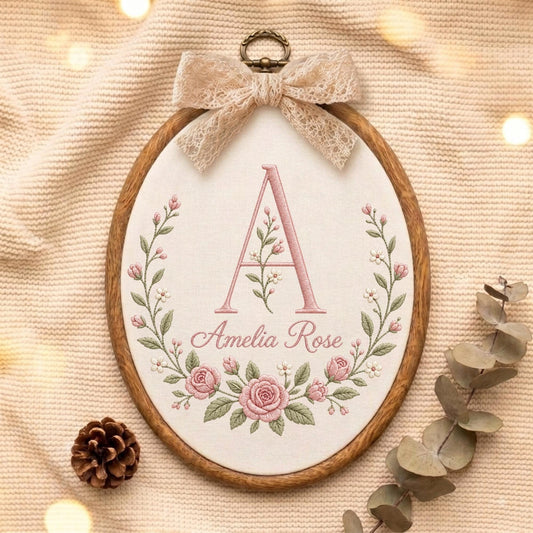 May include: An oval wooden embroidery hoop containing off-white fabric with a floral wreath design in pink and green. The center features a large letter A and the name Amelia Rose in pink script. A beige lace bow is attached to the top.