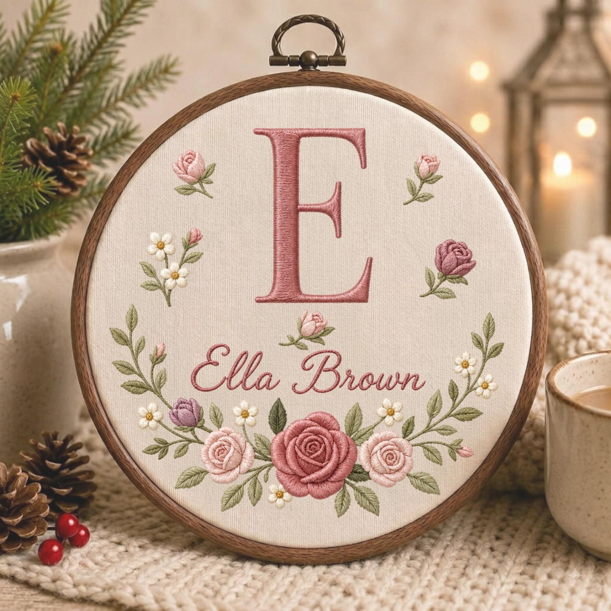 May include: A circular wooden embroidery hoop containing cream-colored fabric with a hand-stitched design. The embroidery features a large pink letter E, the name Ella Brown in cursive script, and a floral wreath of pink roses and small white flowers.