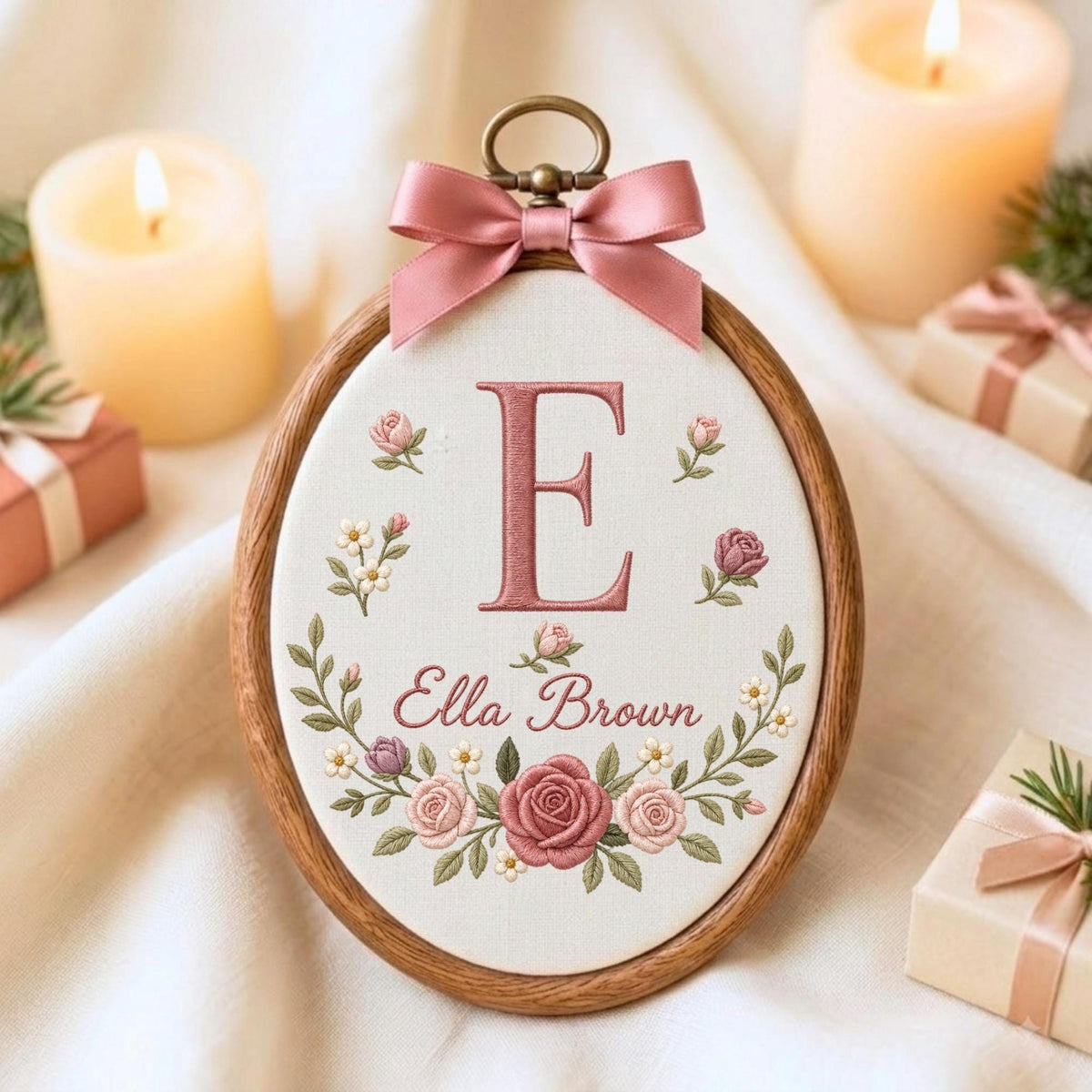 May include: An oval wooden embroidery hoop containing white fabric with a stitched pink letter E and the name Ella Brown in cursive script. The design features embroidered pink roses and small white flowers, topped with a pink satin ribbon bow.