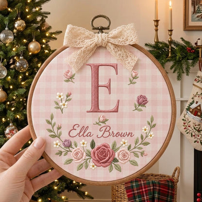May include: A circular wooden embroidery hoop featuring a pink and white gingham fabric background. The design includes a large embroidered letter E in dark pink, the name Ella Brown in cursive, and a floral wreath of roses and small flowers.