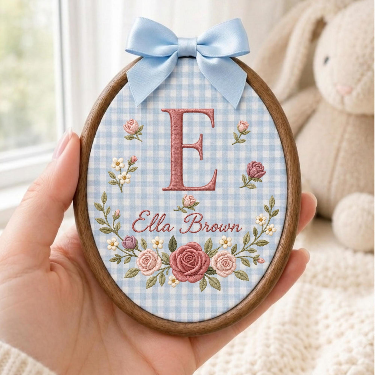 May include: An oval wooden embroidery hoop featuring light blue and white gingham fabric. The center displays a large embroidered letter E in dusty rose, with the name Ella Brown below. A floral wreath of pink roses and small white flowers surrounds the text, topped with a light blue satin ribbon bow.