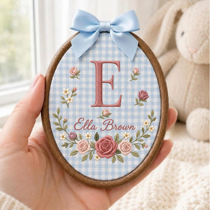 May include: An oval wooden embroidery hoop featuring light blue and white gingham fabric. The center displays a large embroidered letter E in dusty rose, with the name Ella Brown below. A floral wreath of pink roses and small white flowers surrounds the text, topped with a light blue satin ribbon bow.