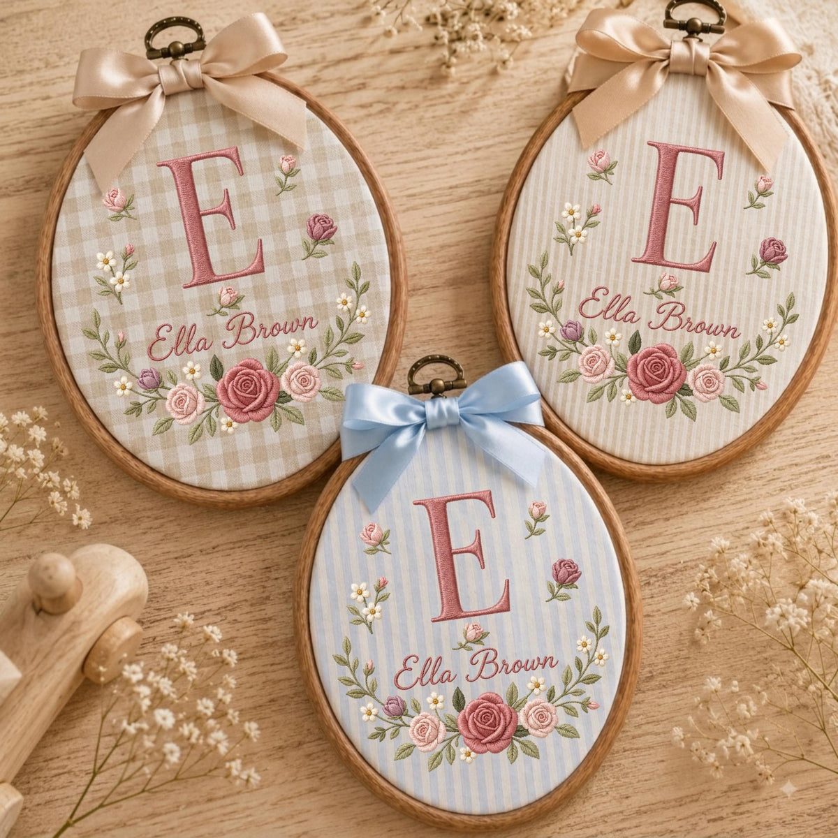 May include: Three oval wooden embroidery hoops featuring fabric backgrounds with the letter E and the name Ella Brown embroidered in pink, surrounded by floral wreaths. Each hoop is topped with a decorative satin ribbon bow in beige or blue.