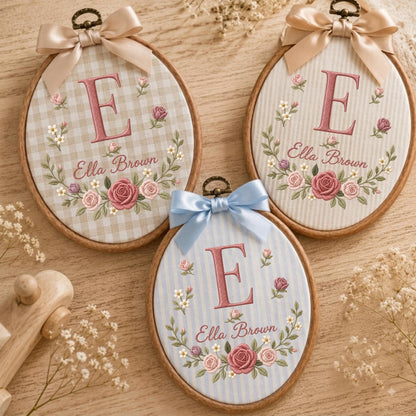 May include: Three oval wooden embroidery hoops featuring fabric backgrounds with the letter E and the name Ella Brown embroidered in pink, surrounded by floral wreaths. Each hoop is topped with a decorative satin ribbon bow in beige or blue.