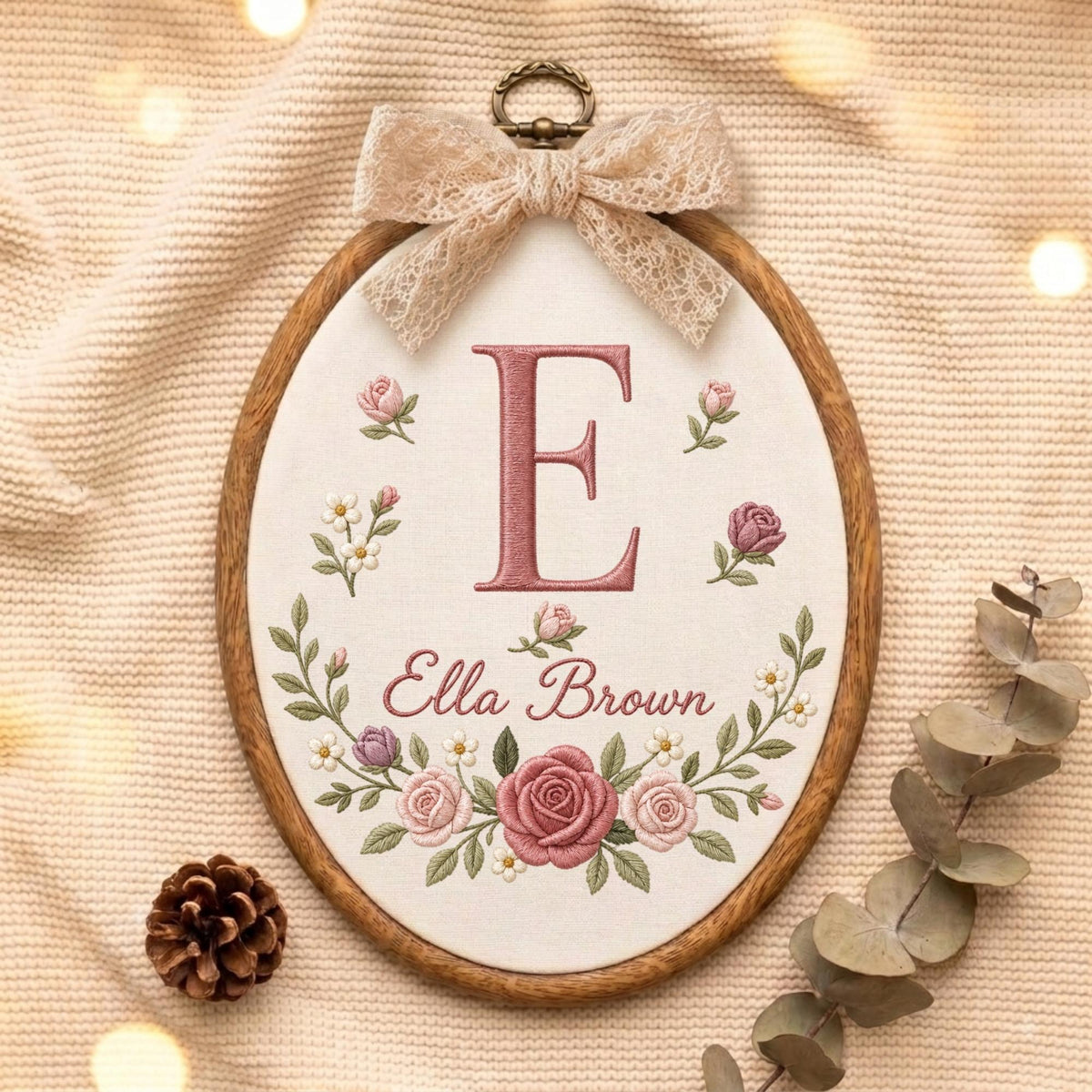 May include: An oval wooden embroidery hoop containing off-white fabric with a large embroidered letter E in dusty rose, the name Ella Brown, and a floral wreath of roses and small white flowers, topped with a beige lace bow and metal hanger.