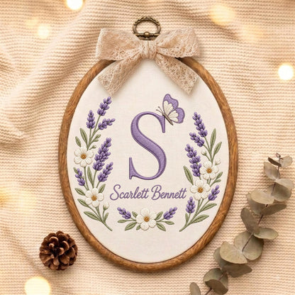 May include: An oval embroidery hoop containing white fabric with a purple embroidered letter S, a butterfly, and lavender floral sprigs. The text Scarlett Bennett is stitched below the letter. A beige lace bow is attached to the top metal hanger.