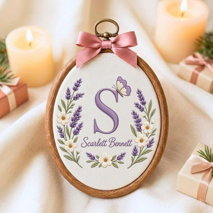 May include: An oval wooden embroidery hoop containing white fabric with a purple embroidered letter S, the name Scarlett Bennett, and a floral wreath of lavender and white flowers with a butterfly, topped with a pink satin ribbon bow.