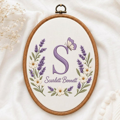 May include: An oval wooden embroidery hoop containing white fabric with a purple embroidered letter S, a purple butterfly, and floral sprigs of lavender and white flowers. The text Scarlett Bennett is embroidered in purple below the letter S.