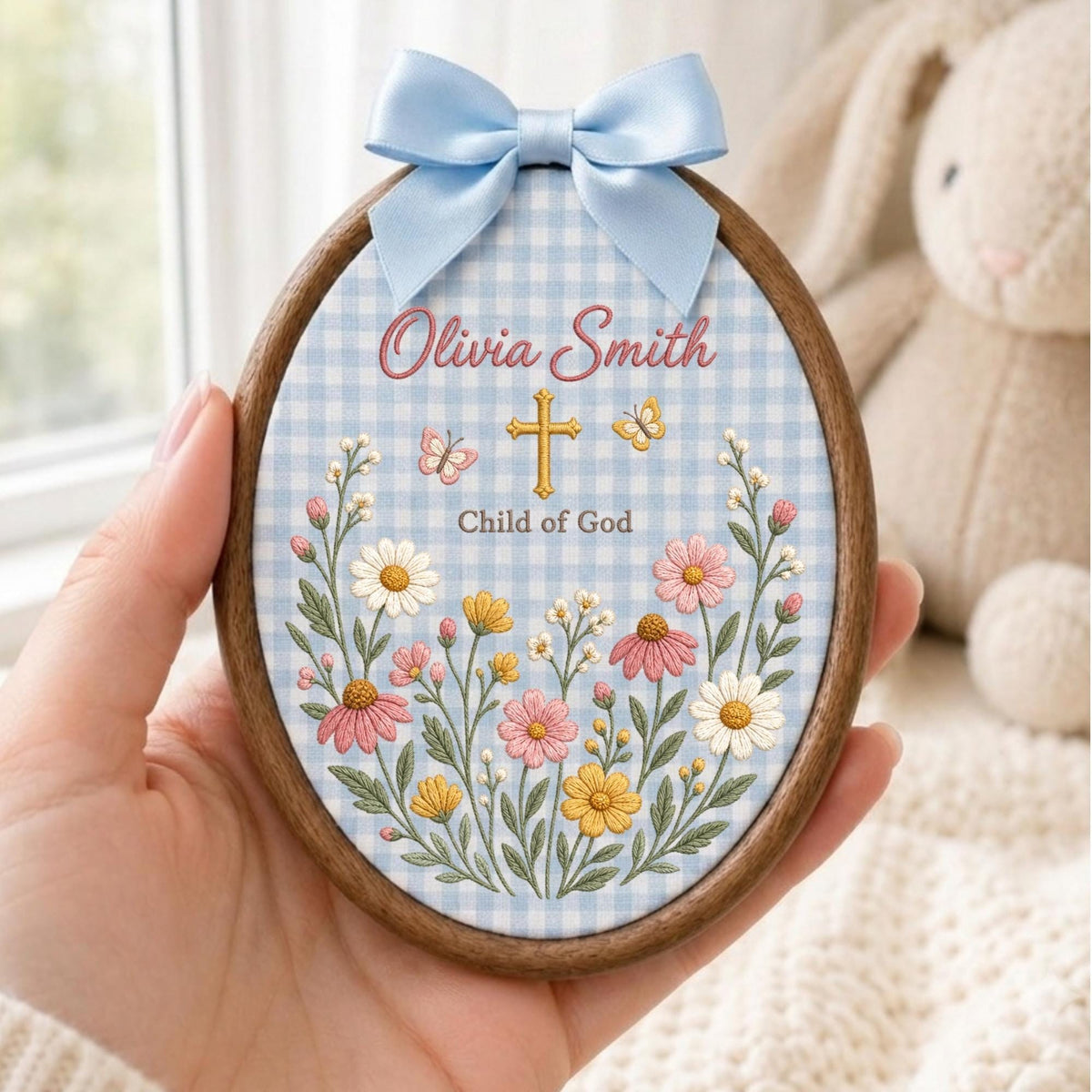 May include: An oval wooden embroidery hoop featuring a light blue and white gingham fabric background. The design includes embroidered pink, yellow, and white wildflowers, a gold cross, two butterflies, and the text Olivia Smith Child of God.
